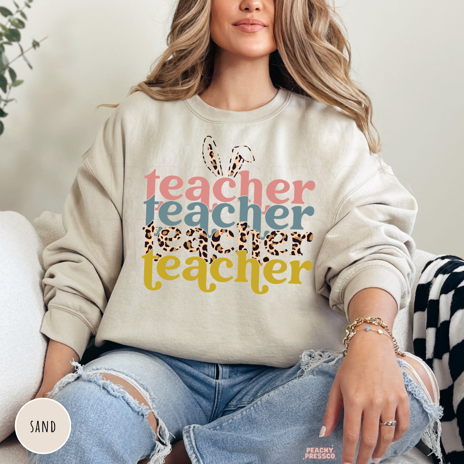 Woman wearing a sand crewneck sweatshirt with a retro Easter Teacher graphic featuring bunny ears, leopard print typography, and pastel colors.