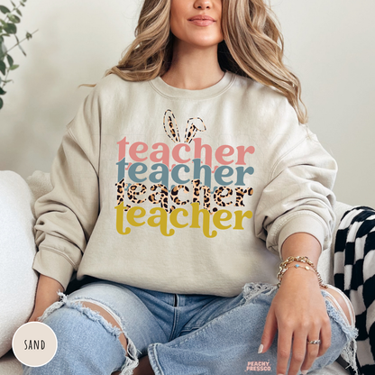 Woman wearing a sand crewneck sweatshirt with a retro Easter Teacher graphic featuring bunny ears, leopard print typography, and pastel colors.