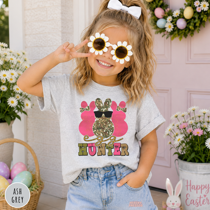 Toddler girl wearing an ash grey Egg Hunter Easter shirt with a pink camo bunny, sunglasses, and festive spring design.