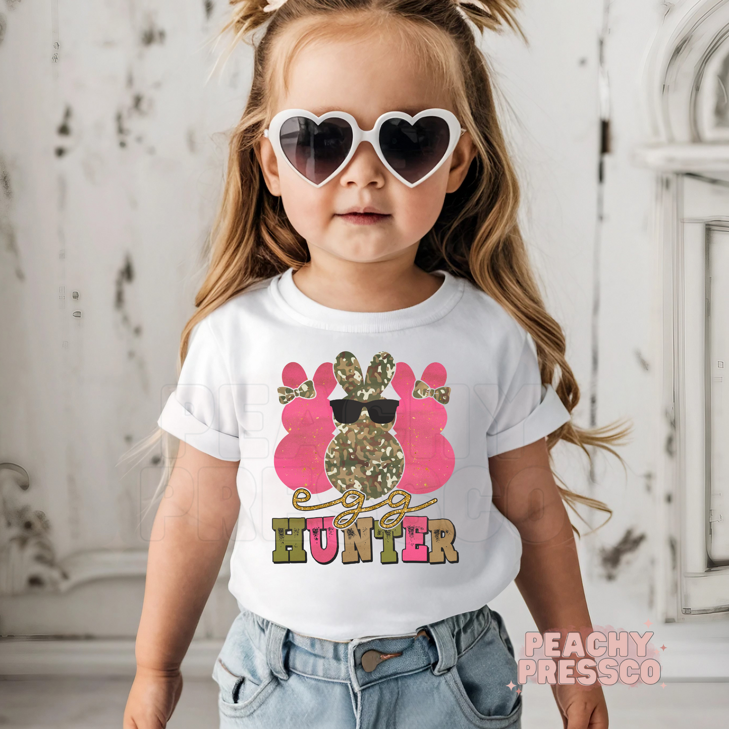 Toddler girl wearing a white Egg Hunter Easter shirt featuring a pink camouflage bunny with sunglasses and a cute spring design.