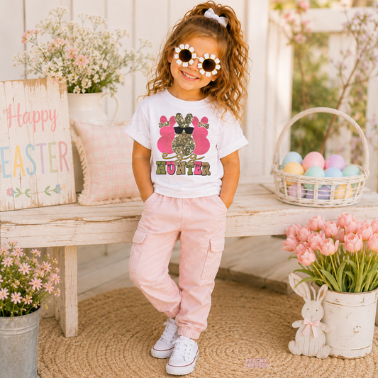 Toddler girl wearing a white Egg Hunter Easter shirt featuring a pink camouflage bunny with sunglasses and a cute spring design.
