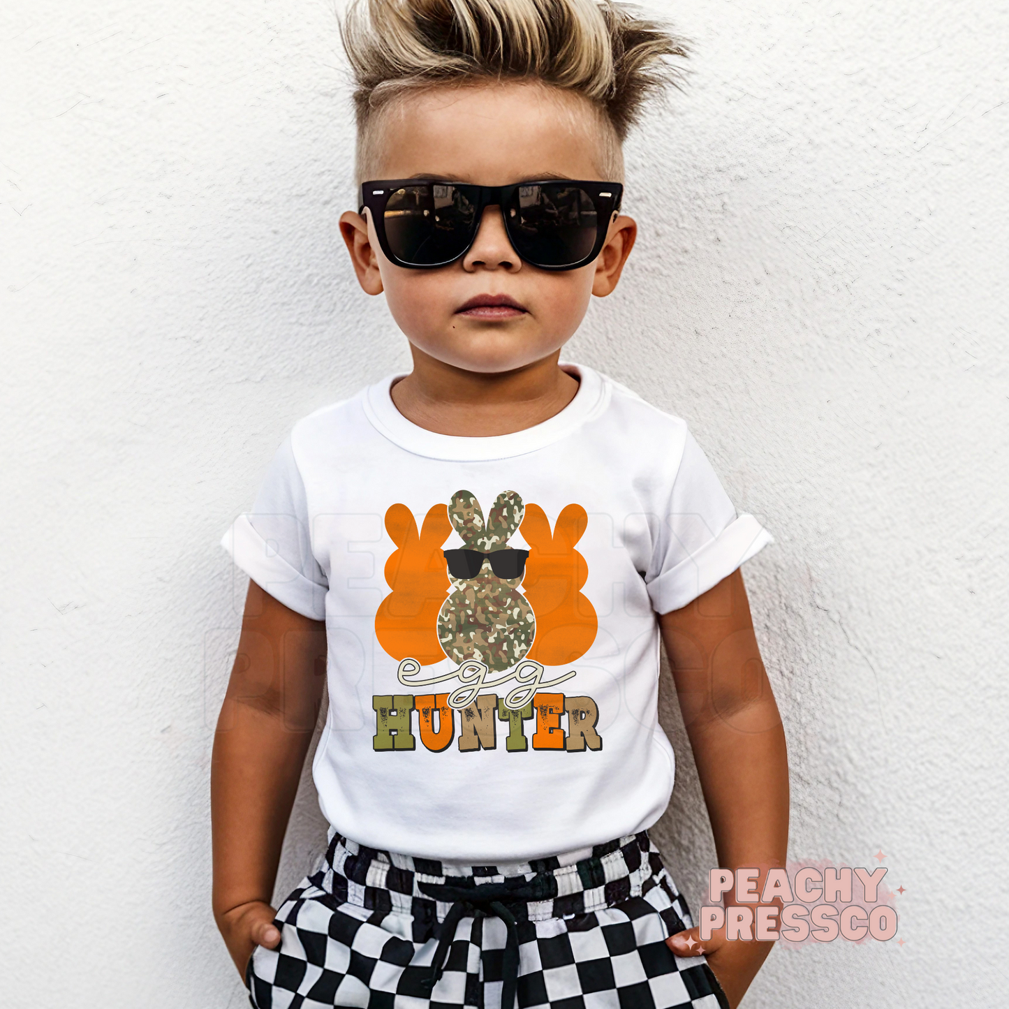 Toddler boy wearing a white Egg Hunter Easter shirt featuring a camouflage bunny with sunglasses and bold orange lettering.