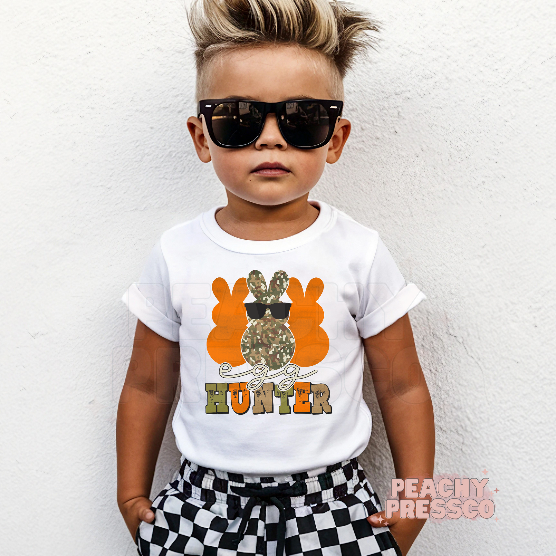 Toddler boy wearing a white Egg Hunter Easter shirt featuring a camouflage bunny with sunglasses and bold orange lettering.