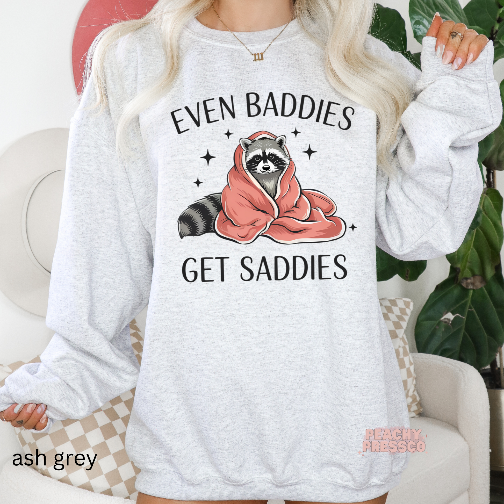 Ash grey crewneck sweatshirt featuring a blanket-wrapped raccoon and the phrase "Even Baddies Get Saddies" in a cute, cozy design.
