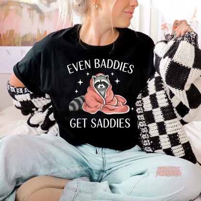 Black graphic t-shirt featuring a cute raccoon wrapped in a blanket with the phrase "Even Baddies Get Saddies."