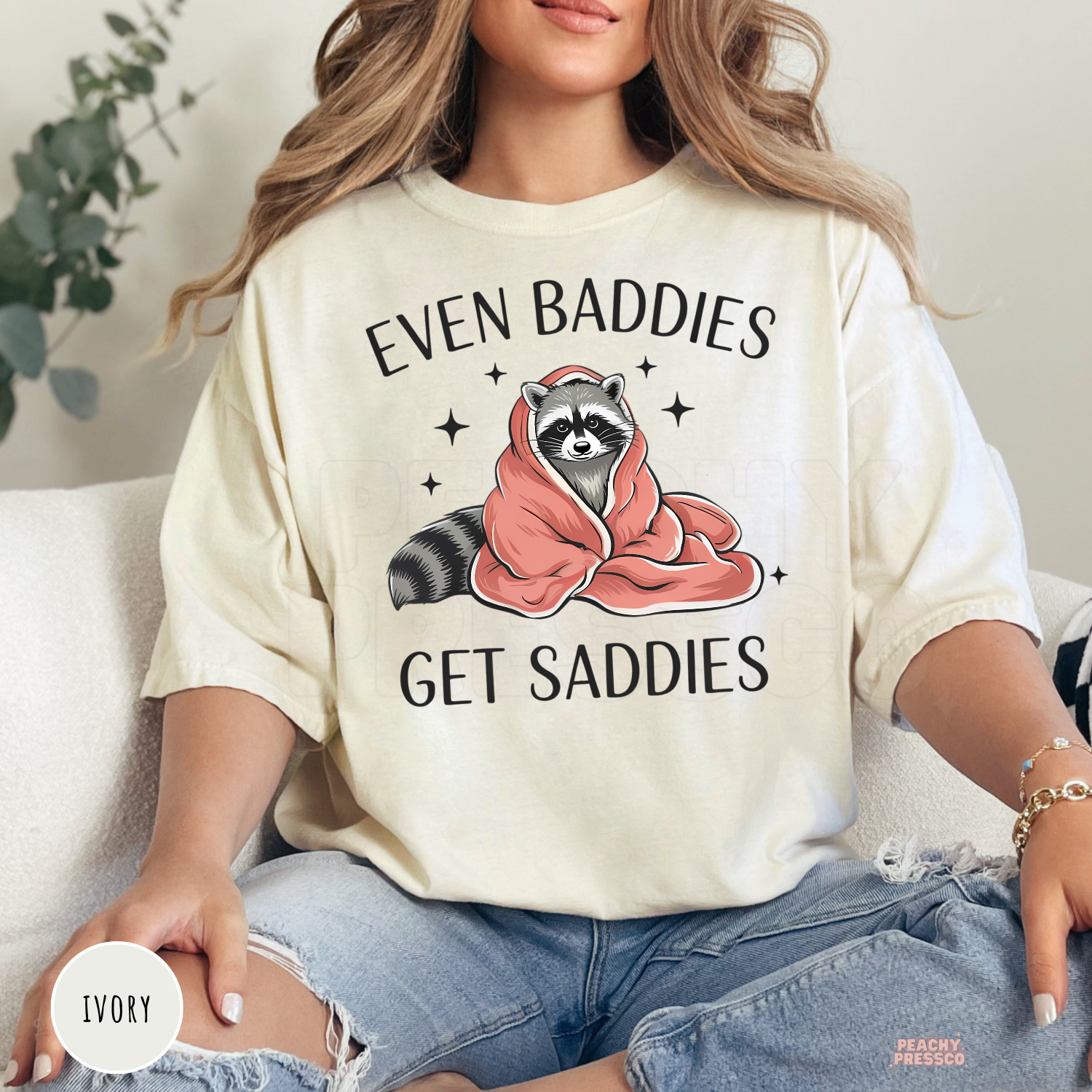 Ivory Comfort Colors t-shirt featuring a cozy raccoon illustration and the funny mental health-inspired quote "Even Baddies Get Saddies."
