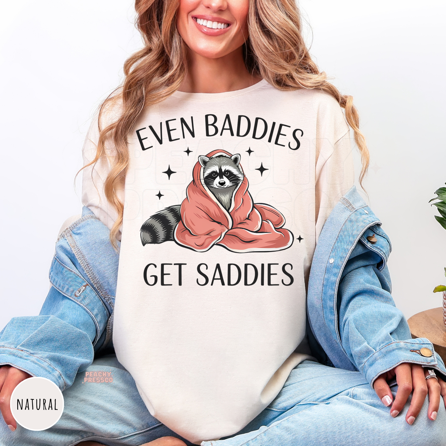 Natural shirt with a cute raccoon wrapped in a blanket and the phrase "Even Baddies Get Saddies."