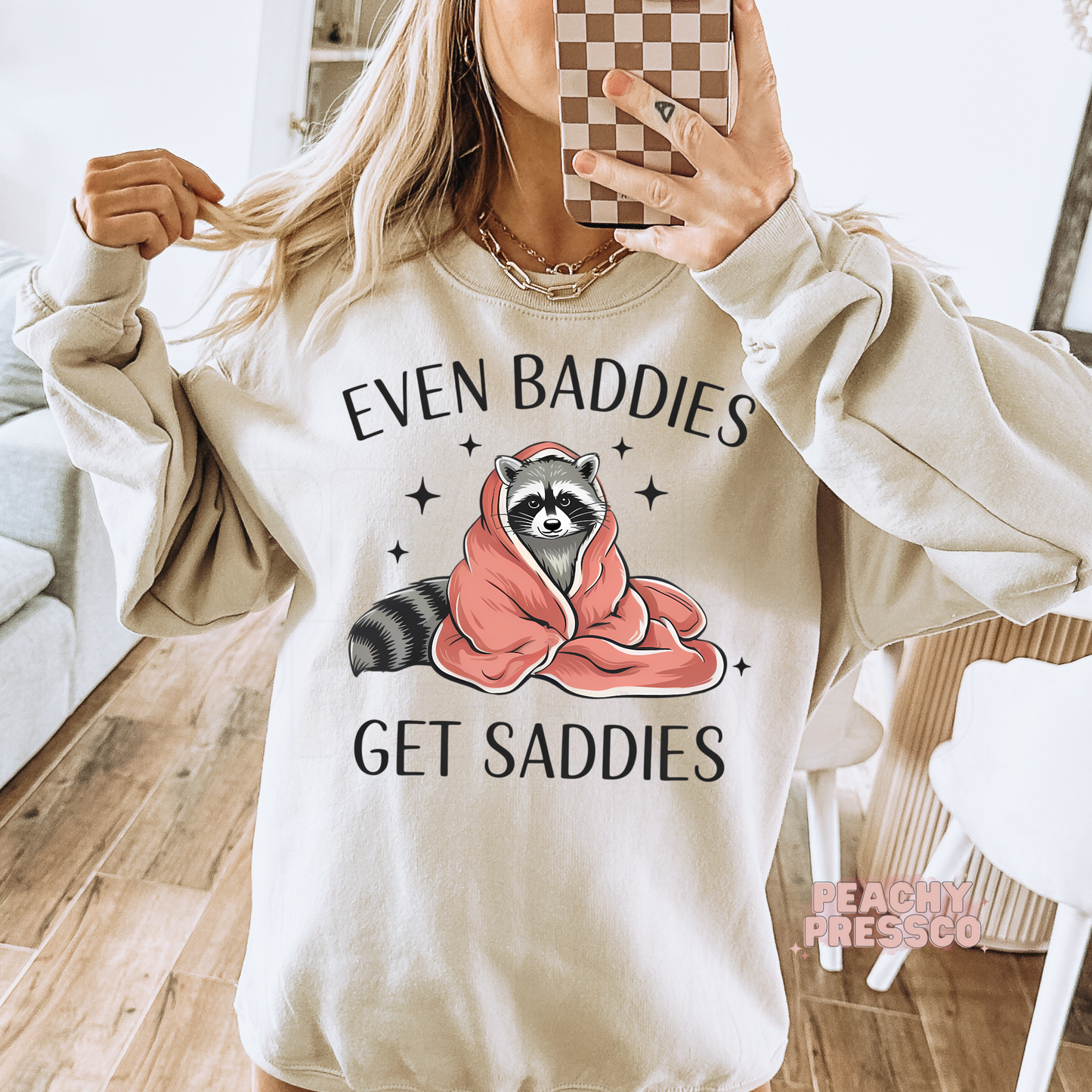Sand crewneck sweatshirt displayed in a mirror selfie featuring a cozy raccoon wrapped in a blanket with the quote "Even Baddies Get Saddies."