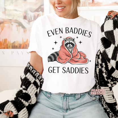 White unisex graphic t-shirt with a cozy raccoon illustration and the funny quote "Even Baddies Get Saddies."