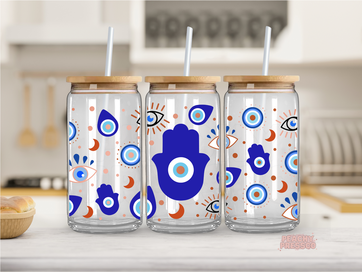 Clear glass cup with a bamboo lid and reusable straw featuring an all-over evil eye and Hamsa hand design in blue, peach, and white. Boho glass tumbler perfect for iced coffee, tea, and everyday drinks.
