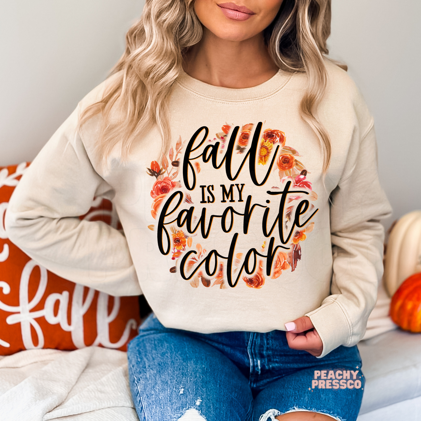 Woman wearing a sand crewneck sweatshirt featuring a floral autumn "Fall Is My Favorite Color" graphic in warm seasonal colors.