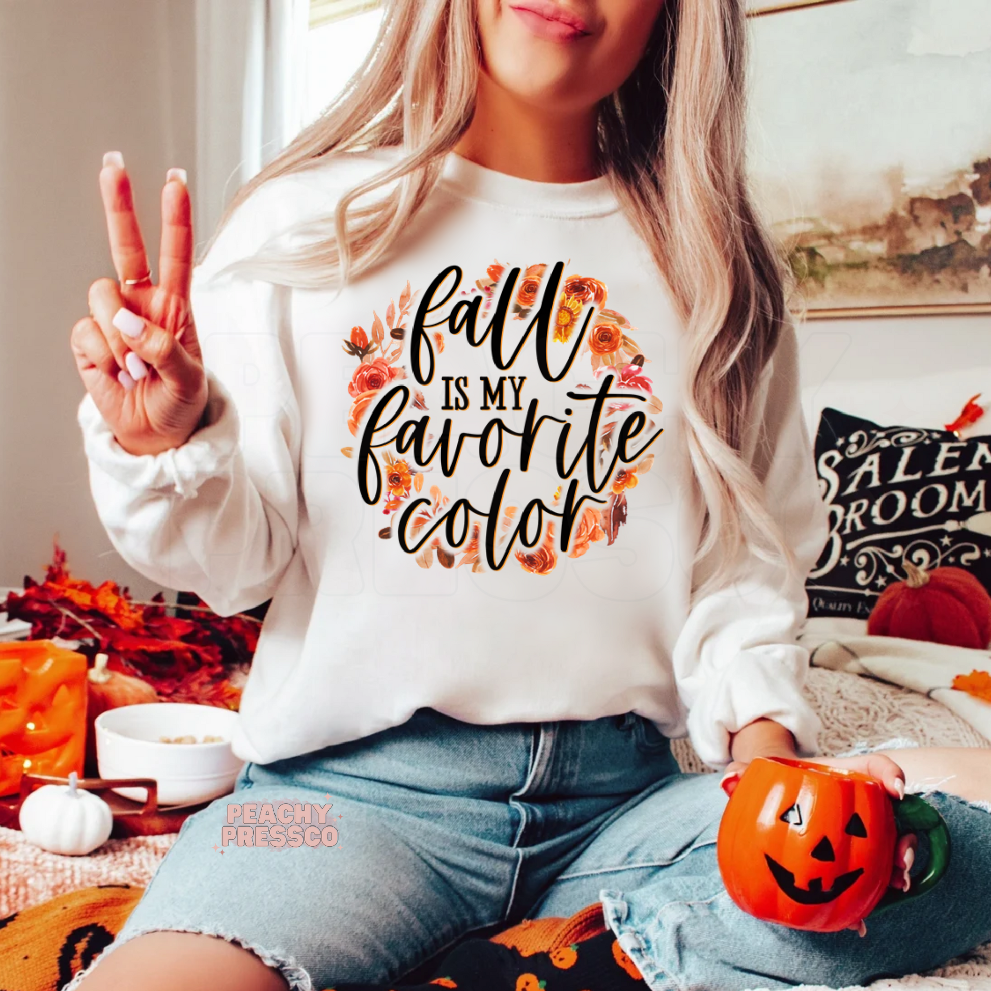 Woman wearing a white crewneck sweatshirt with a "Fall Is My Favorite Color" floral autumn graphic while holding a pumpkin mug.