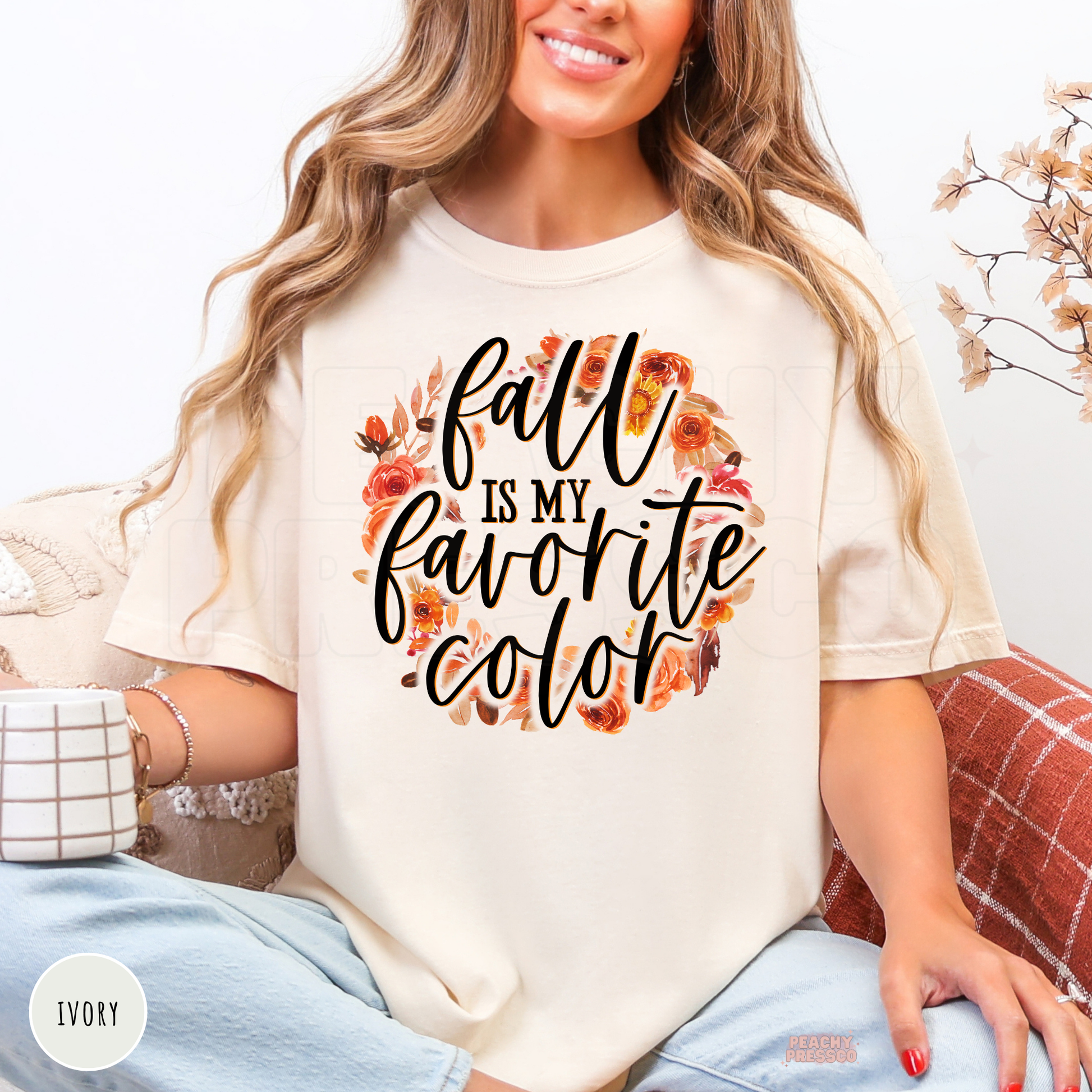 Woman wearing an ivory graphic t-shirt featuring the phrase "Fall Is My Favorite Color" with a floral autumn wreath in orange and neutral tones.