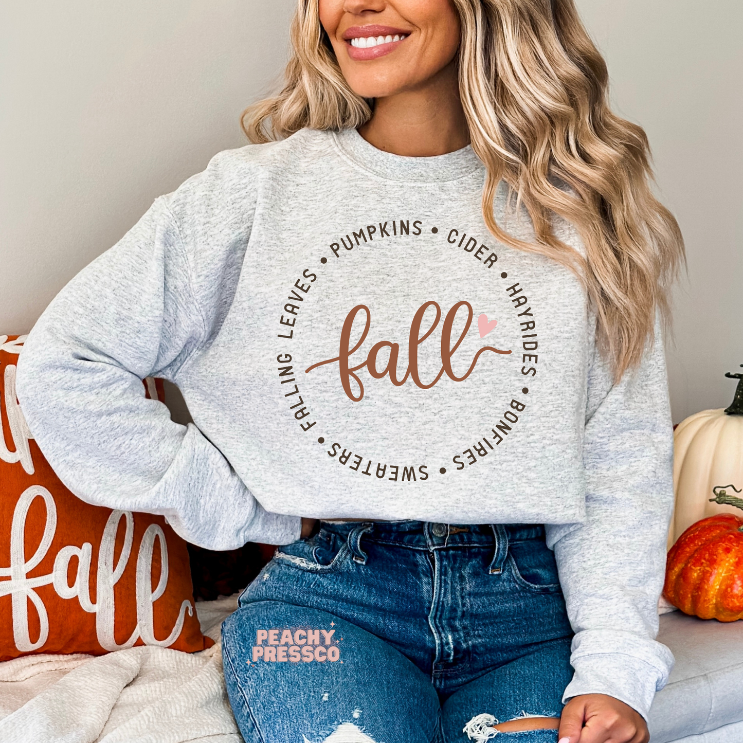 Woman wearing an ash grey crewneck sweatshirt with a modern circular fall graphic featuring pumpkins, cider, hayrides, bonfires, sweaters, and falling leaves.