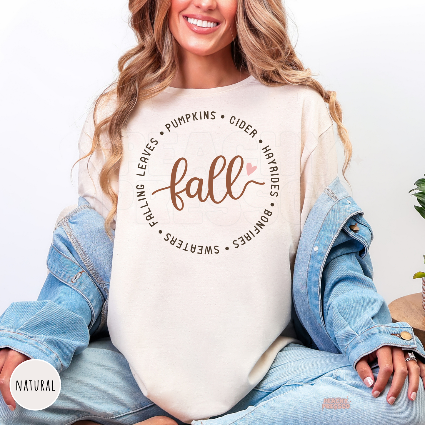 Woman wearing a natural shirt featuring a minimalist fall design with the word "Fall" surrounded by pumpkins, cider, hayrides, bonfires, falling leaves, and sweaters.