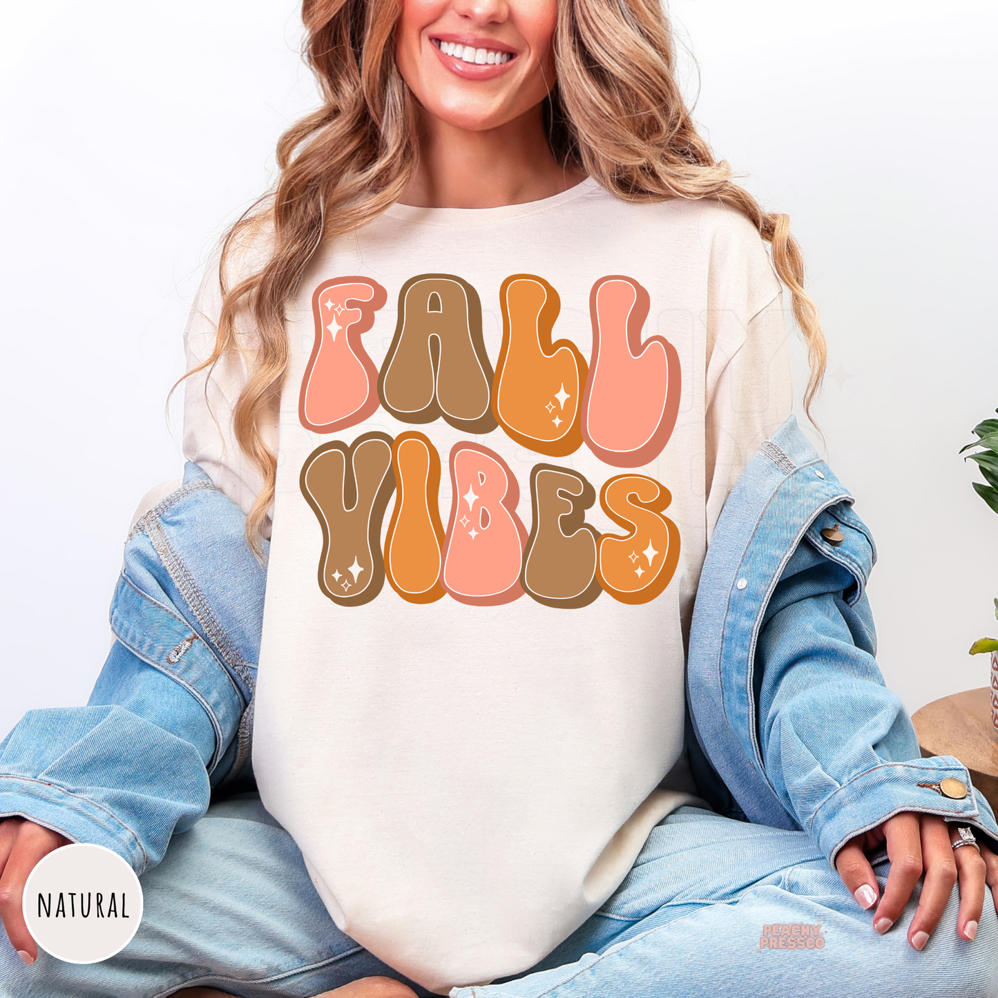 Woman wearing a natural graphic t-shirt featuring a retro "Fall Vibes" design in pink, orange, and brown bubble lettering for the autumn season.