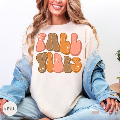 Woman wearing a natural graphic t-shirt featuring a retro "Fall Vibes" design in pink, orange, and brown bubble lettering for the autumn season.