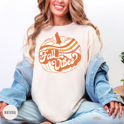 Woman wearing a natural graphic t-shirt featuring a vintage Fall Vibes pumpkin design in warm retro autumn colors.