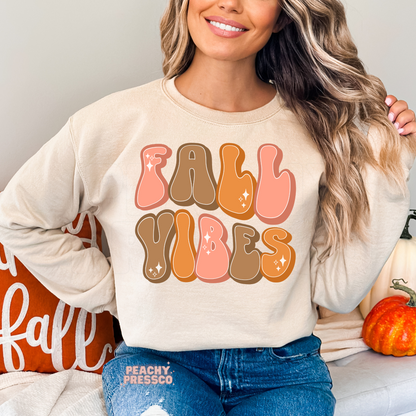 Woman wearing a sand crewneck sweatshirt with a colorful retro "Fall Vibes" typography design in warm autumn tones.