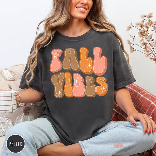 Woman wearing a pepper Comfort Colors t-shirt featuring a bold retro "Fall Vibes" graphic in pink, orange, and brown bubble lettering.
