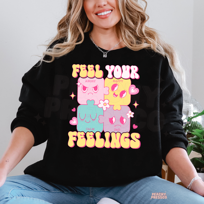 Black Feel Your Feelings sweatshirt featuring colourful puzzle characters representing happy, sad, angry and calm moods with vibrant pink and yellow lettering.