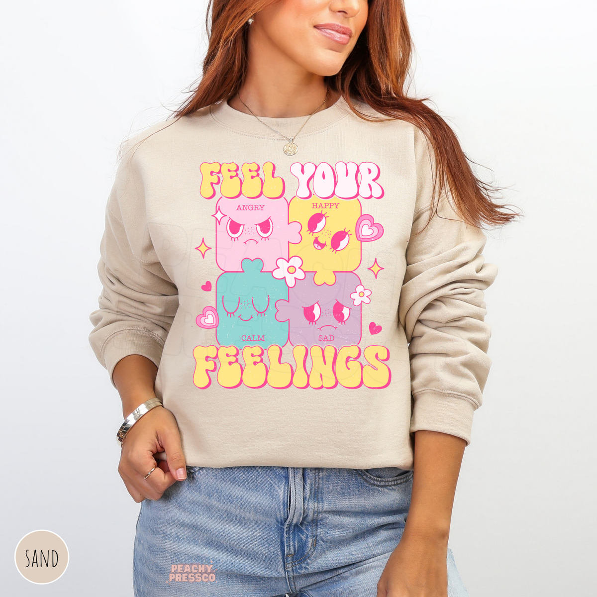 Sand Feel Your Feelings sweatshirt with playful pastel emotion characters, pink hearts, sparkling stars and bright yellow retro typography promoting emotional awareness.