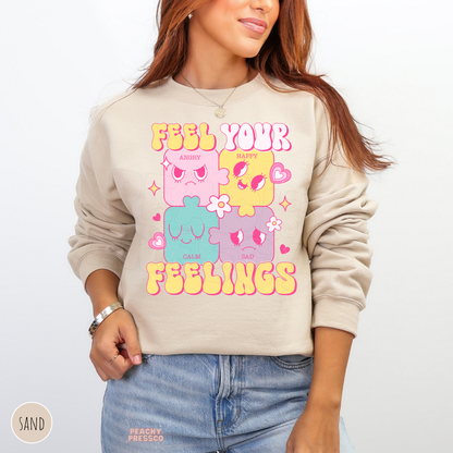 Sand Feel Your Feelings sweatshirt with playful pastel emotion characters, pink hearts, sparkling stars and bright yellow retro typography promoting emotional awareness.