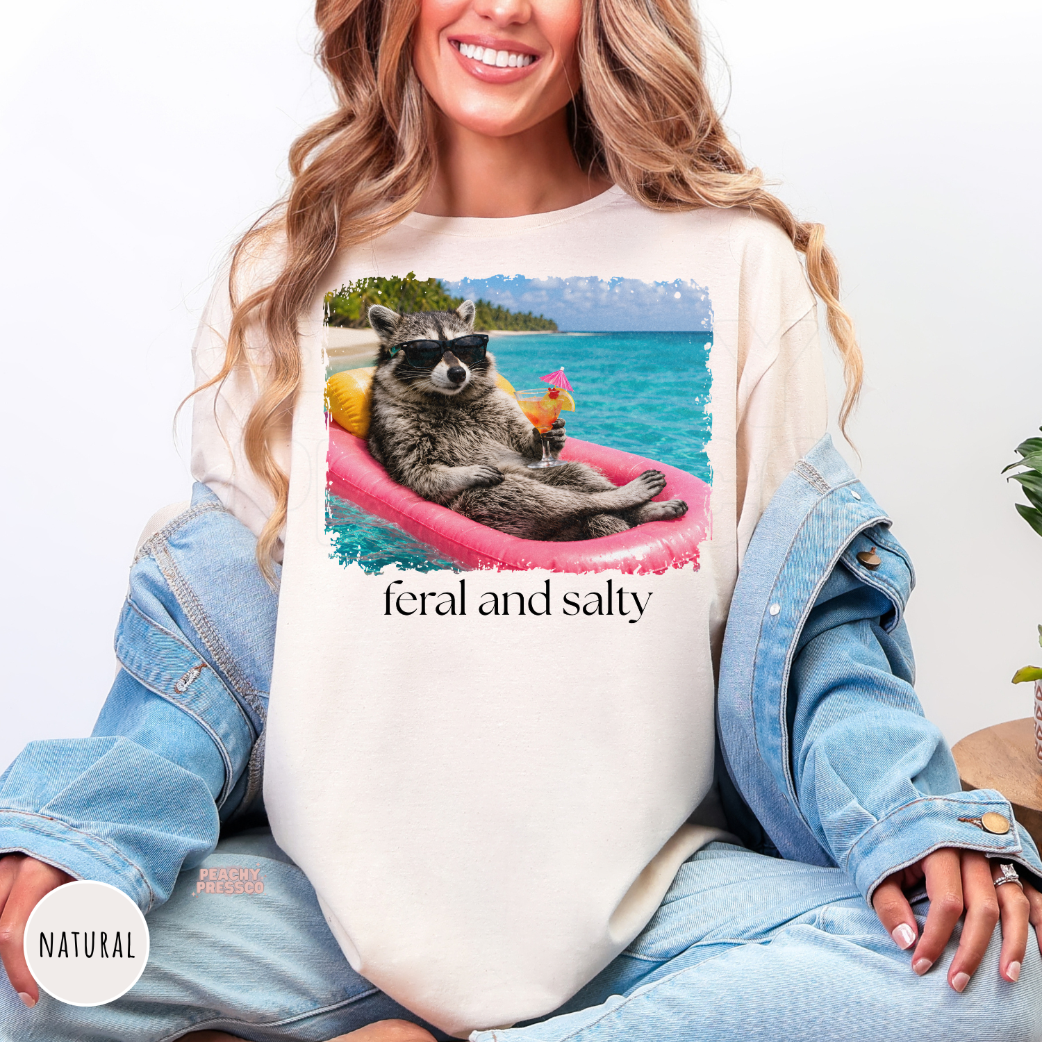 Natural unisex t-shirt featuring a funny "Feral and Salty" raccoon relaxing on a pink pool float with a tropical drink at the beach. Perfect beach vacation, lake days, and summer lovers.