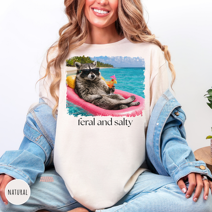 Natural unisex t-shirt featuring a funny "Feral and Salty" raccoon relaxing on a pink pool float with a tropical drink at the beach. Perfect beach vacation, lake days, and summer lovers.
