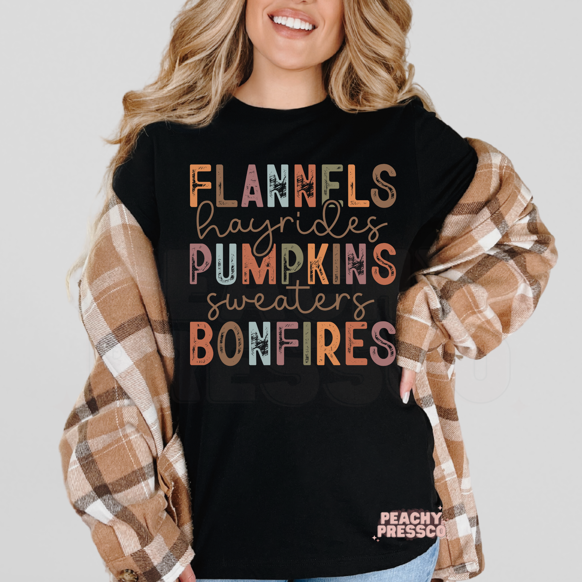 Woman wearing a black graphic t-shirt featuring the phrase "Flannels Hayrides Pumpkins Sweaters Bonfires" in colorful vintage fall lettering.