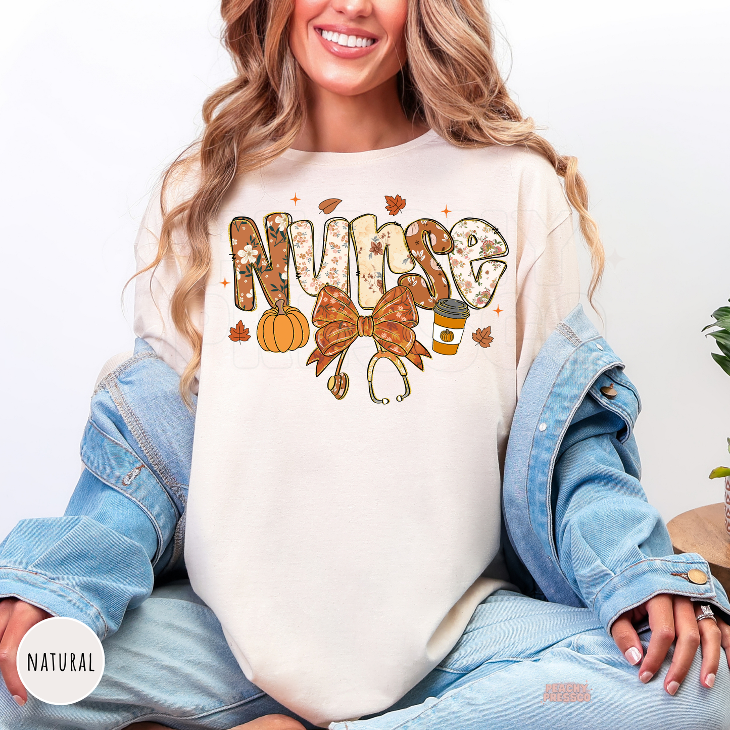 Woman wearing a natural graphic t-shirt featuring a floral "Nurse" fall design with a pumpkin, coffee cup, stethoscope bow, and autumn leaves.