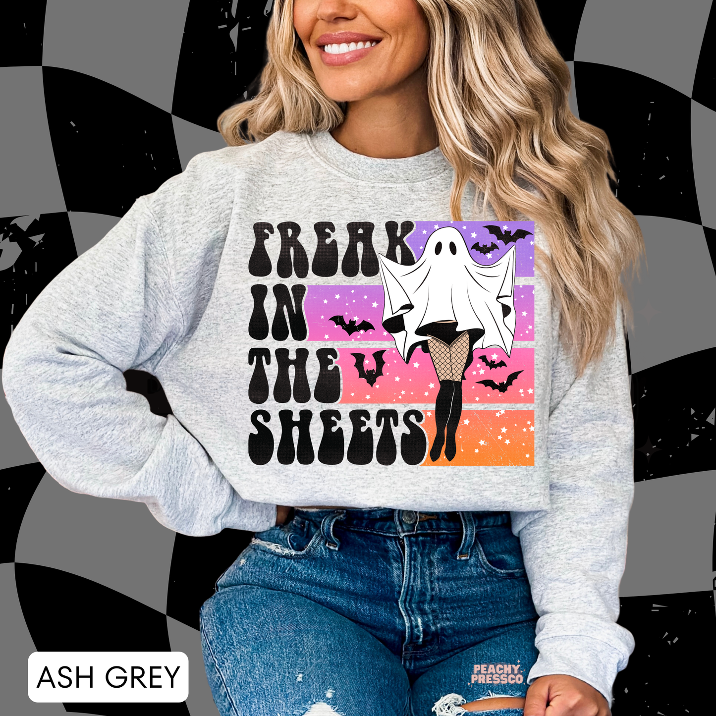 Woman wearing an ash grey Halloween crewneck sweatshirt with a Freak in the Sheets ghost graphic, bats and colorful retro sunset.