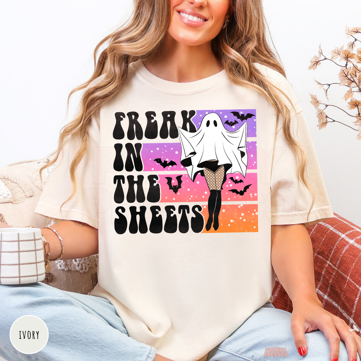 Woman wearing an ivory Comfort Colors Halloween t-shirt featuring a funny "Freak in the Sheets" ghost graphic with fishnet stockings, bats and retro sunset.