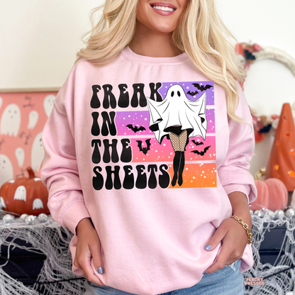 Woman wearing a light pink Halloween sweatshirt featuring a funny Freak in the Sheets ghost graphic with retro sunset, bats and spooky season aesthetic.