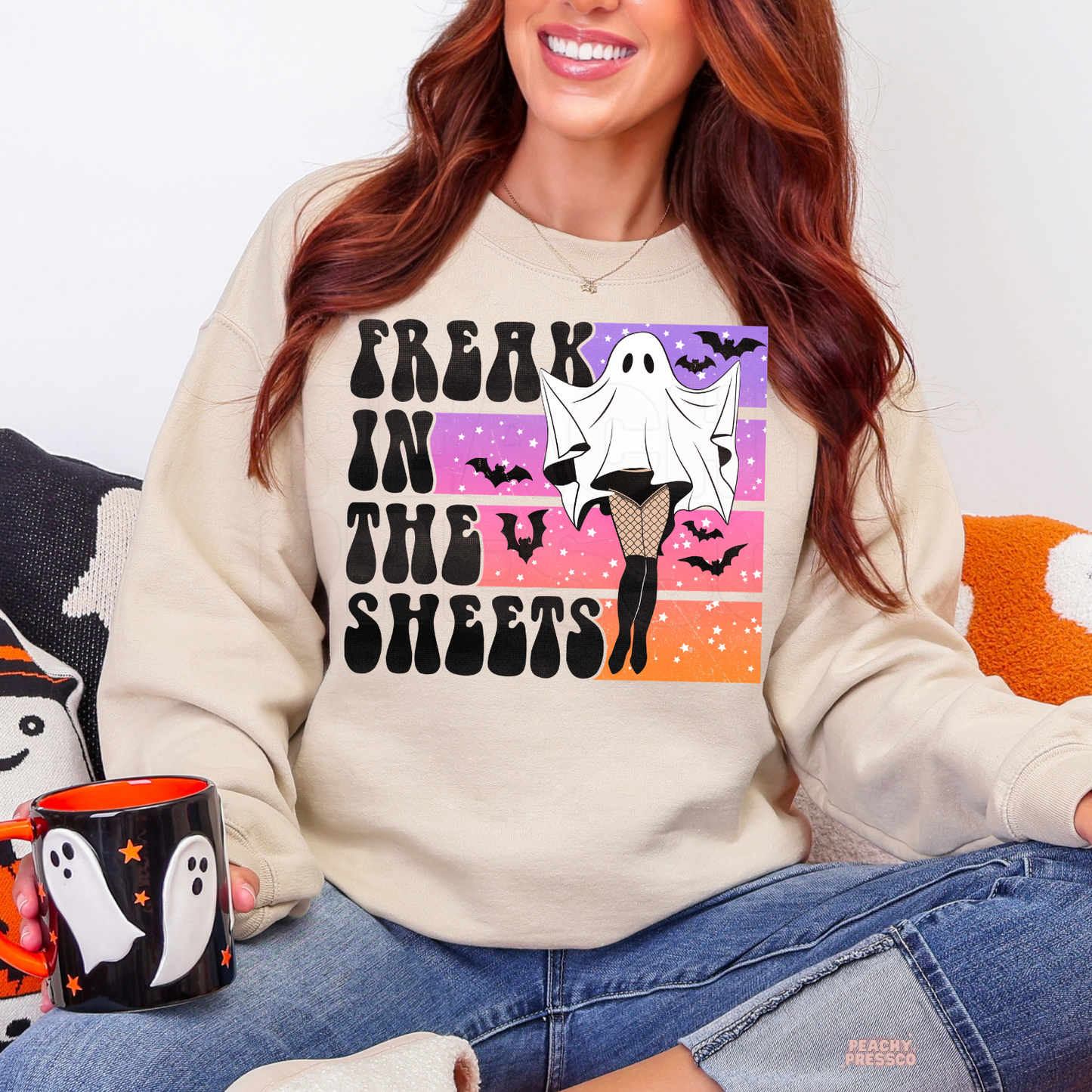 Woman wearing a sand Halloween crewneck sweatshirt featuring a funny Freak in the Sheets ghost graphic with retro sunset colors, bats and fishnet stockings while holding a Halloween mug.