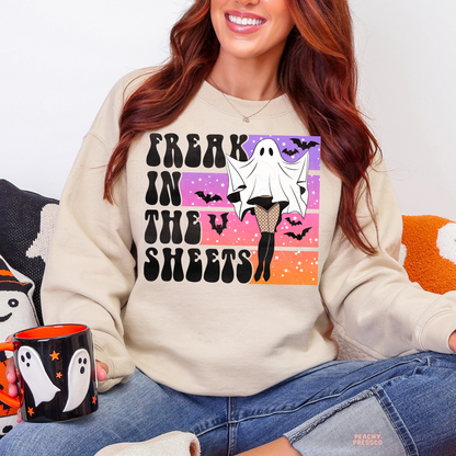 Woman wearing a sand Halloween crewneck sweatshirt featuring a funny Freak in the Sheets ghost graphic with retro sunset colors, bats and fishnet stockings while holding a Halloween mug.