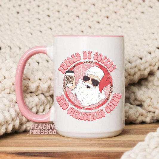 Ceramic Christmas mug with pink handle featuring Santa Claus wearing sunglasses holding a leopard print coffee cup with text Fueled By Coffee And Christmas Cheer.