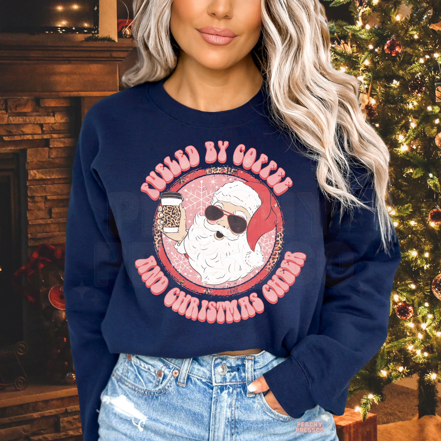 Woman standing in front of a Christmas tree wearing a navy blue crewneck sweatshirt featuring a cool sunglasses-wearing Santa and coffee print.