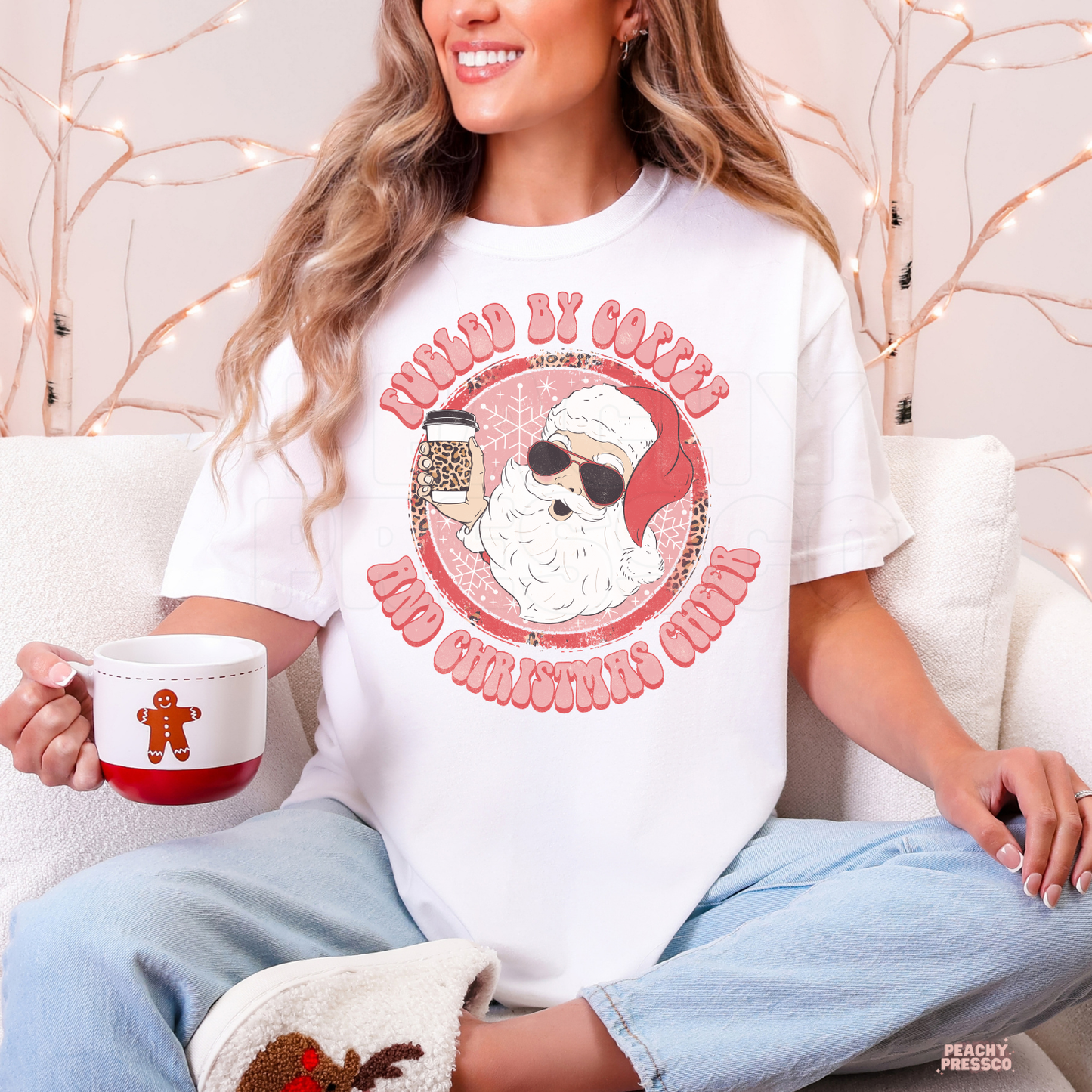Woman holding a gingerbread mug sitting on a couch wearing a white t-shirt printed with a retro Santa holding a coffee cup.