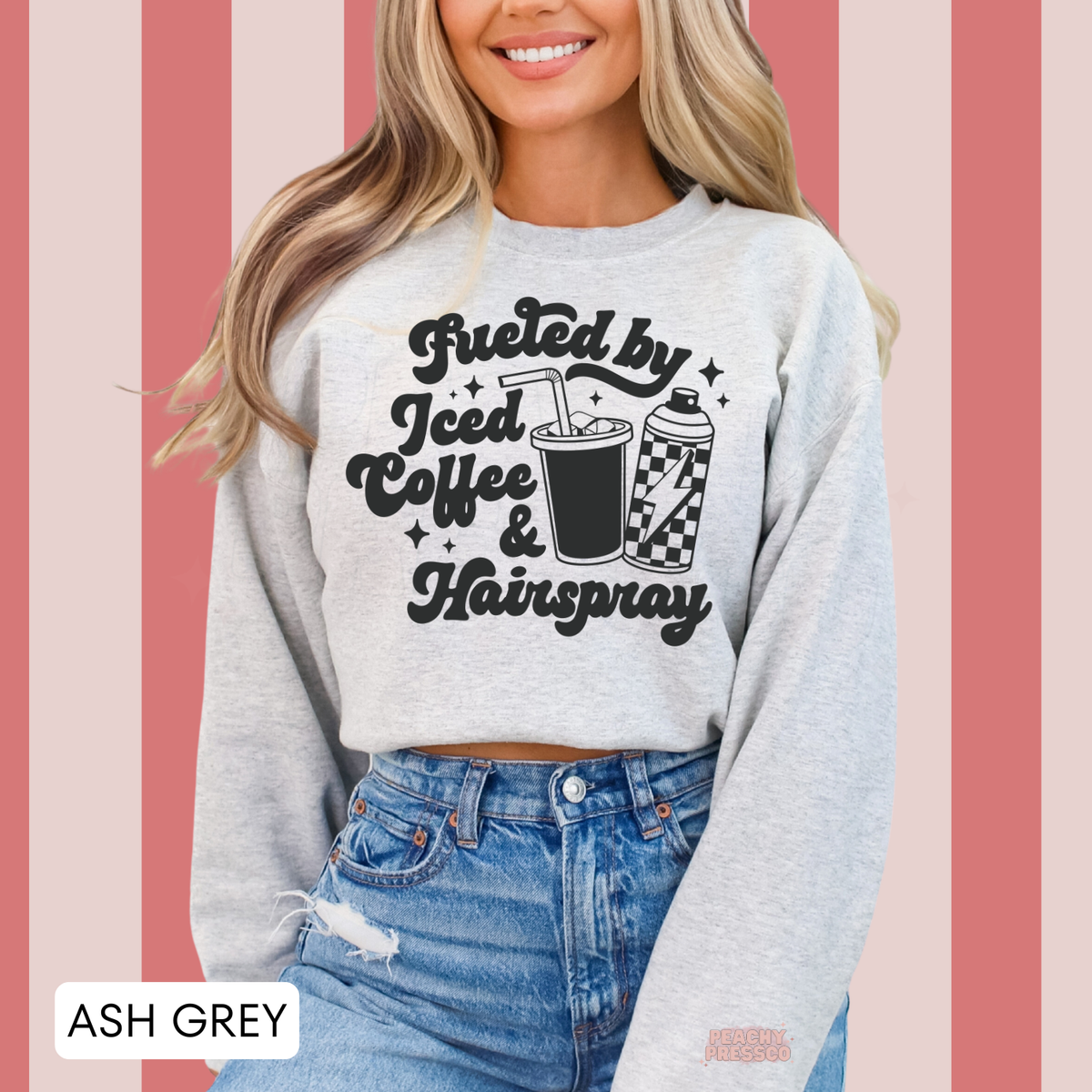 Woman wearing an ash grey Fueled by Iced Coffee & Hairspray sweatshirt featuring black retro lettering, iced coffee and hairspray illustrations against a pink striped background.
