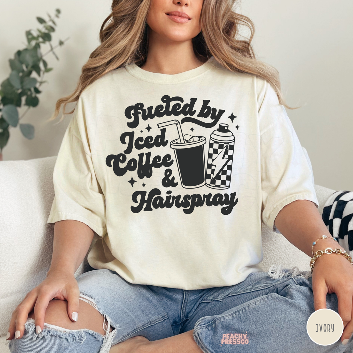 Woman wearing an ivory Fueled by Iced Coffee & Hairspray hairstylist T-shirt with black retro lettering, iced coffee cup, checkered hairspray can and sparkles.