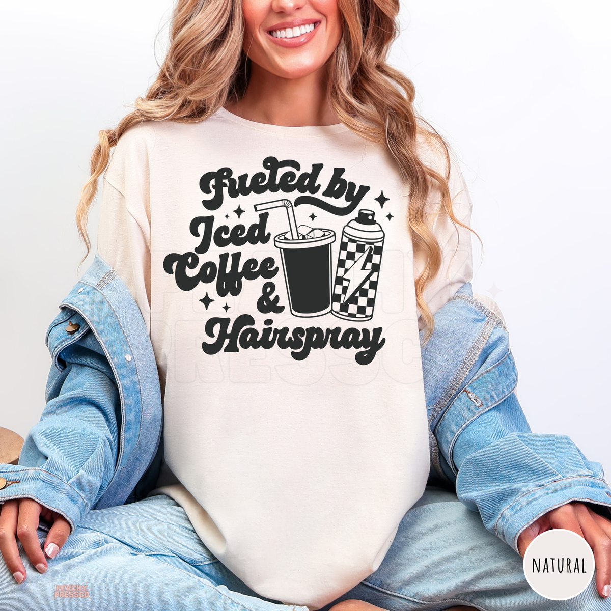 Woman wearing a natural colour Fueled by Iced Coffee & Hairspray hairdresser T-shirt with bold retro typography, iced coffee illustration and hairspray can.