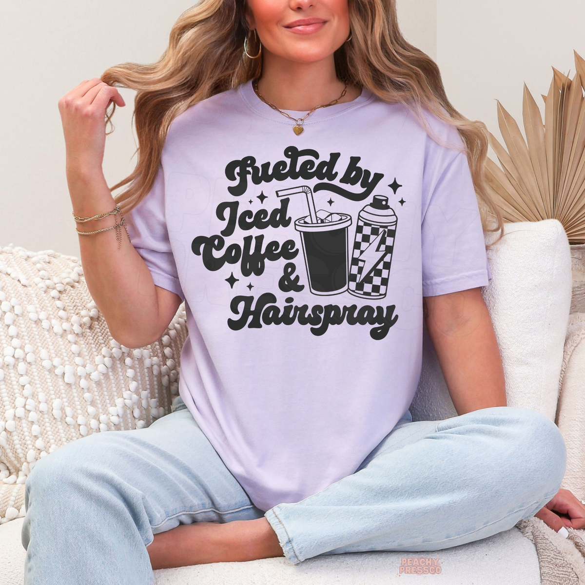Woman wearing a orchid Fueled by Iced Coffee & Hairspray hairstylist T-shirt with black lettering, iced coffee cup, checkered hairspray can and star accents.