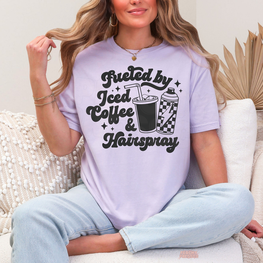 Woman wearing a orchid Fueled by Iced Coffee & Hairspray hairstylist T-shirt with black lettering, iced coffee cup, checkered hairspray can and star accents.