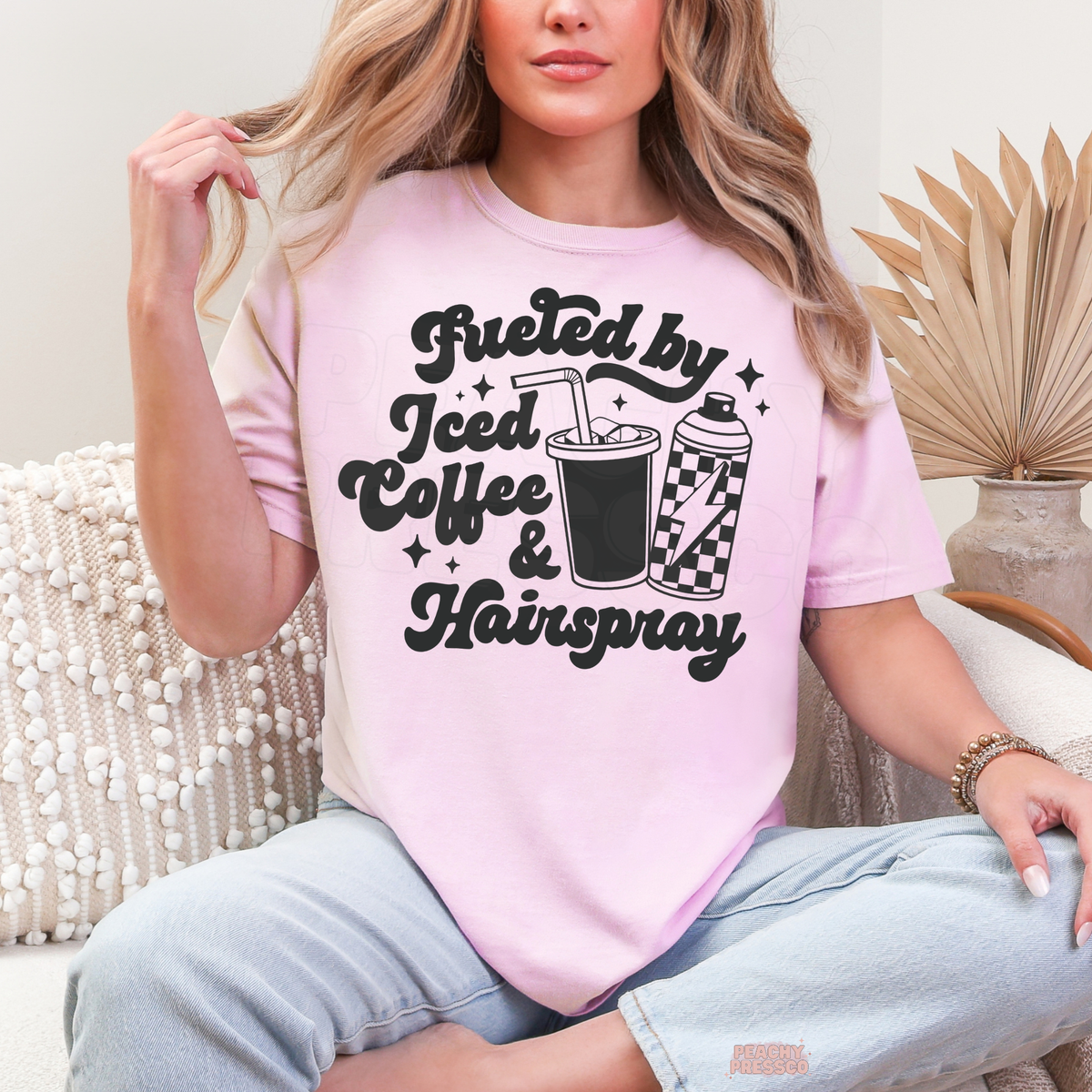 Woman wearing a light pink Fueled by Iced Coffee & Hairspray T-shirt featuring bold black typography, iced coffee with straw, checkered hairspray can and stars.