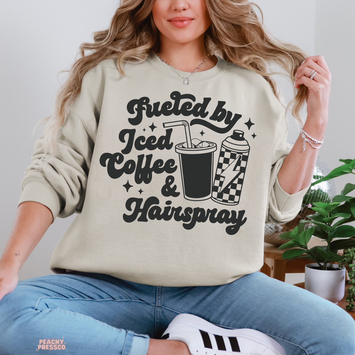 Woman wearing a sand Fueled by Iced Coffee & Hairspray crewneck sweatshirt featuring bold black lettering, iced coffee and checkered hairspray graphics.