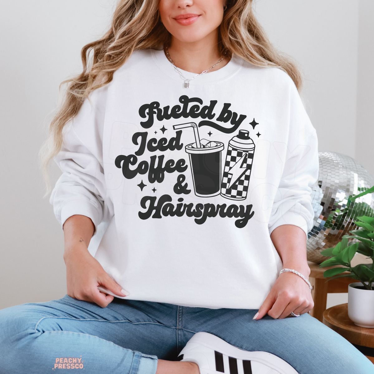 Woman wearing a white Fueled by Iced Coffee & Hairspray hairstylist sweatshirt with black retro lettering, iced coffee cup, checkered hairspray can and sparkles.