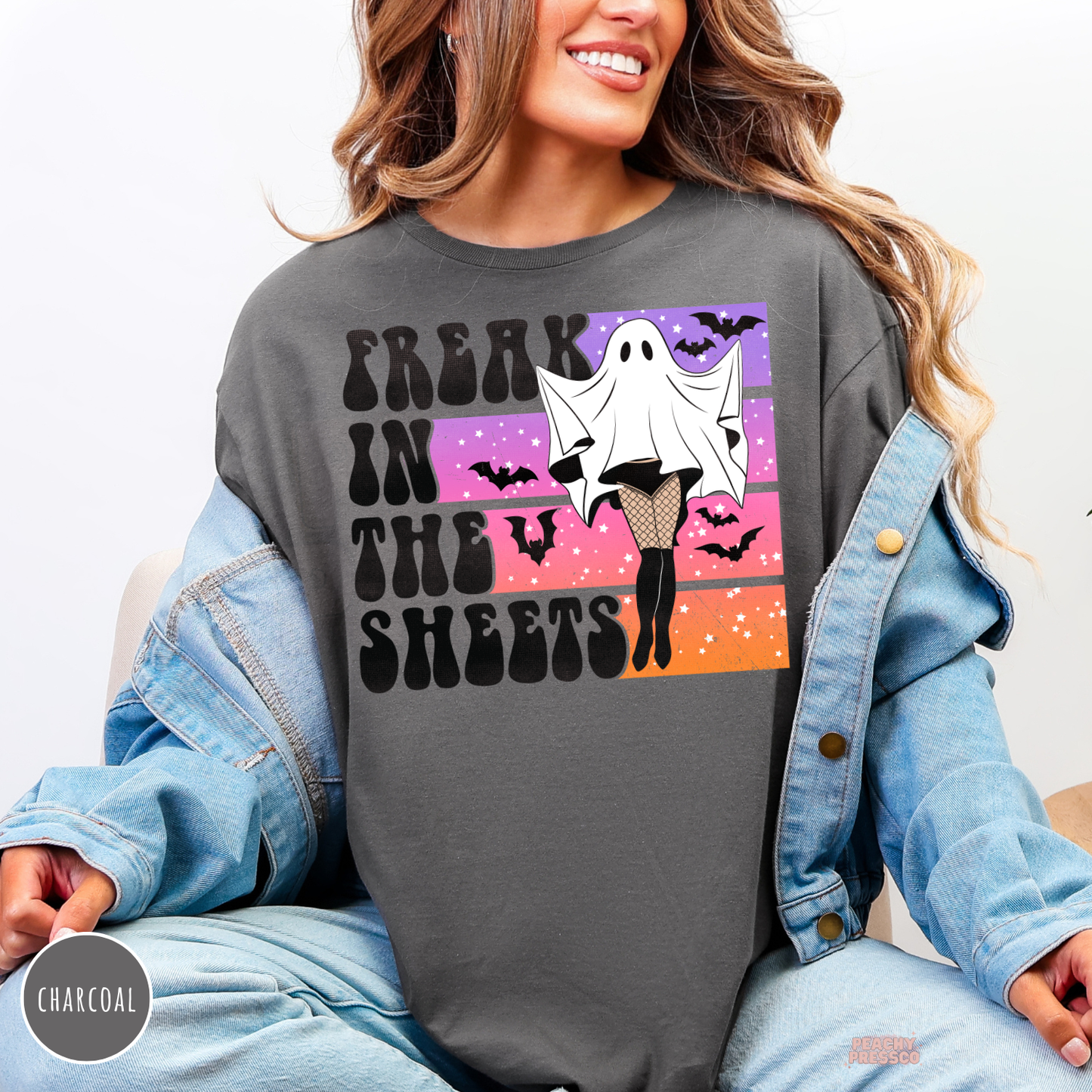 Woman wearing a charcoal Halloween crewneck sweatshirt with a funny Freak in the Sheets ghost graphic, retro sunset, bats and spooky vintage design.