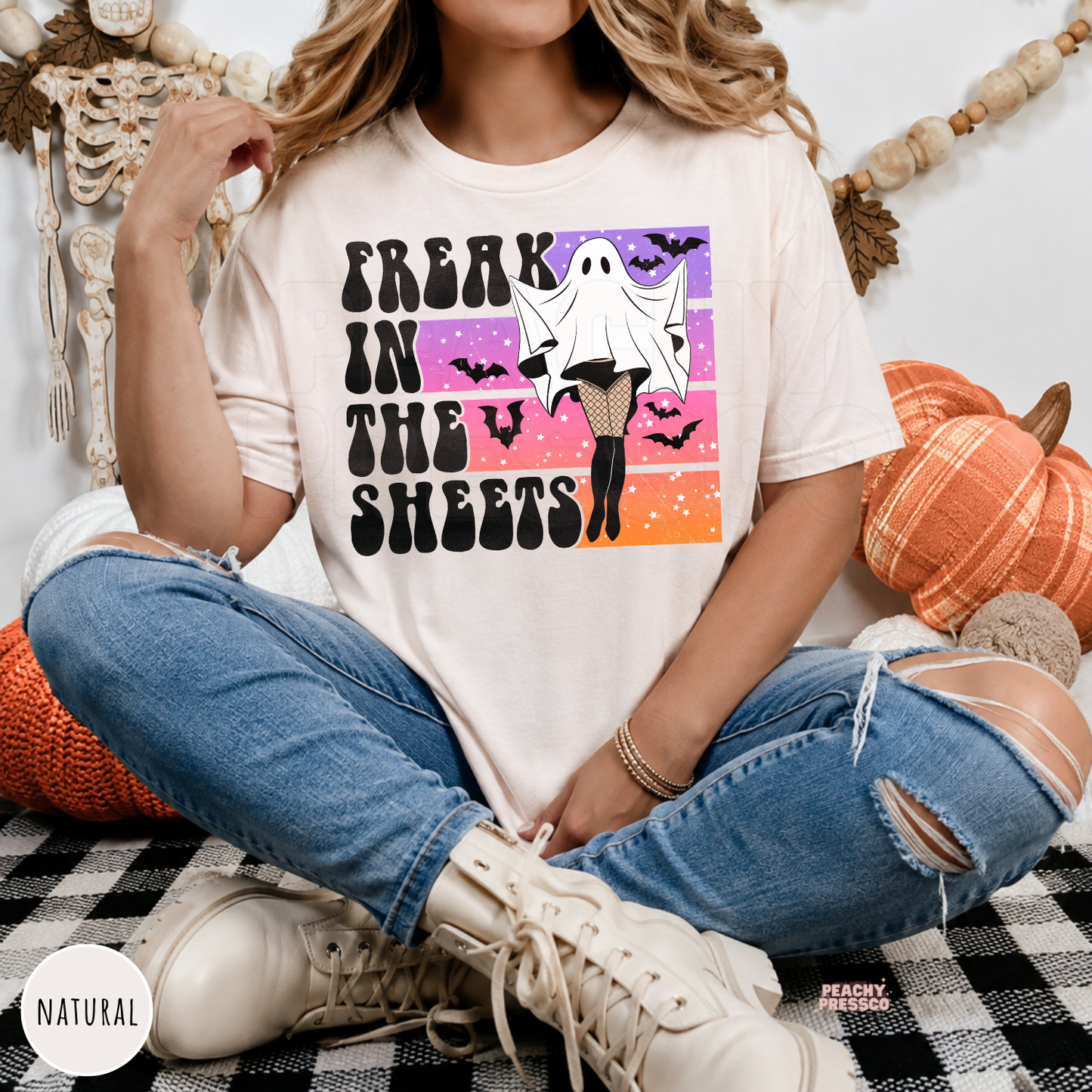Woman wearing a natural Halloween graphic tee with a funny Freak in the Sheets ghost design, bats and retro vintage sunset artwork.