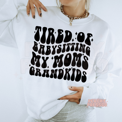 White sweatshirt with the funny "Tired of Babysitting My Mom's Grandkids" graphic in bold retro typography. Cute motherhood sweatshirt for moms with a sense of humor.